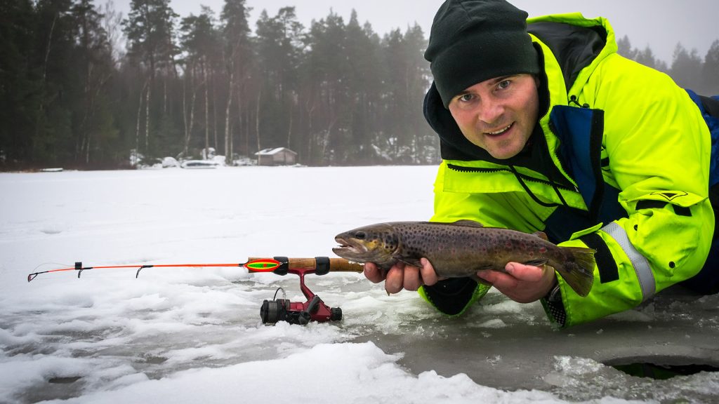 A man in a bright yellow jacket shows off a fish on the ice, surrounded by a winter landscape.